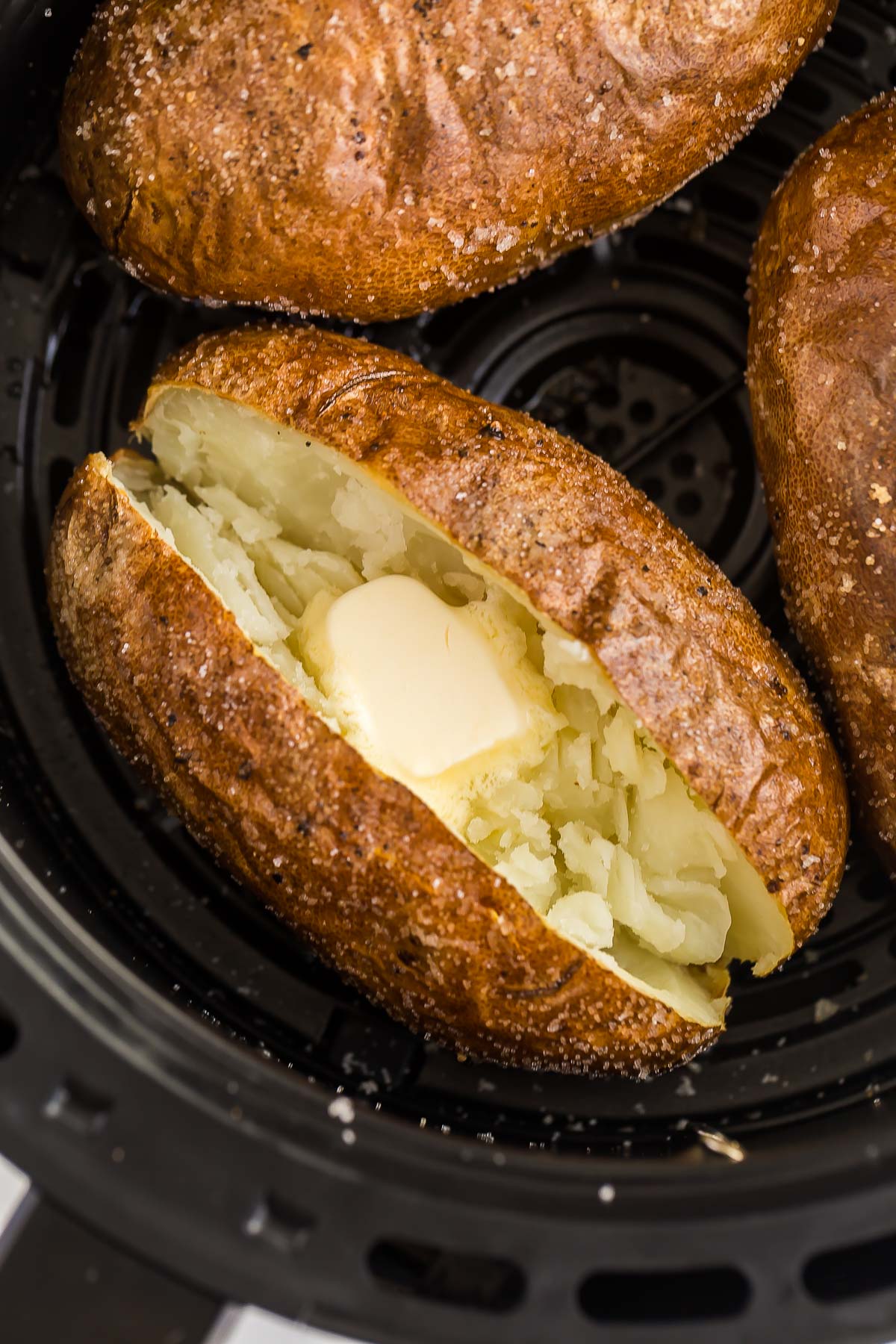 This Air Fryer Baked Potato recipe is incredibly crispy on the outside, fluffy and creamy on the inside, and couldn't be easier! Simply seasoned and perfect for your favorite toppings. #airfryer #airfryerrecipes #potato #recipes | air fryer recipes | air fryer potatoes | crispy baked potato | russet potatoes baked potato in air fryer basket cut open with butter.