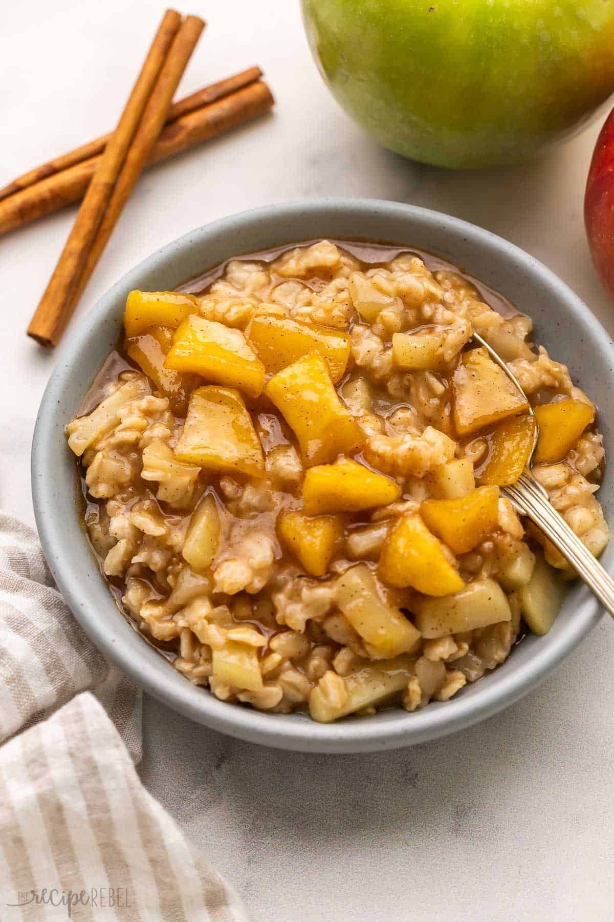 This Apple Cinnamon Oatmeal recipe is packed with chunks of fresh apples swirled throughout warm creamy oats and topped with maple syrup. It's ready in 20 minutes and is gluten-free, dairy-free, and meal prep-friendly! #breakfast #recipe #apples | apple oatmeal | oatmeal recipe | how to cook oatmeal | apple recipes grey bowl filled with oatmeal and apples beside.