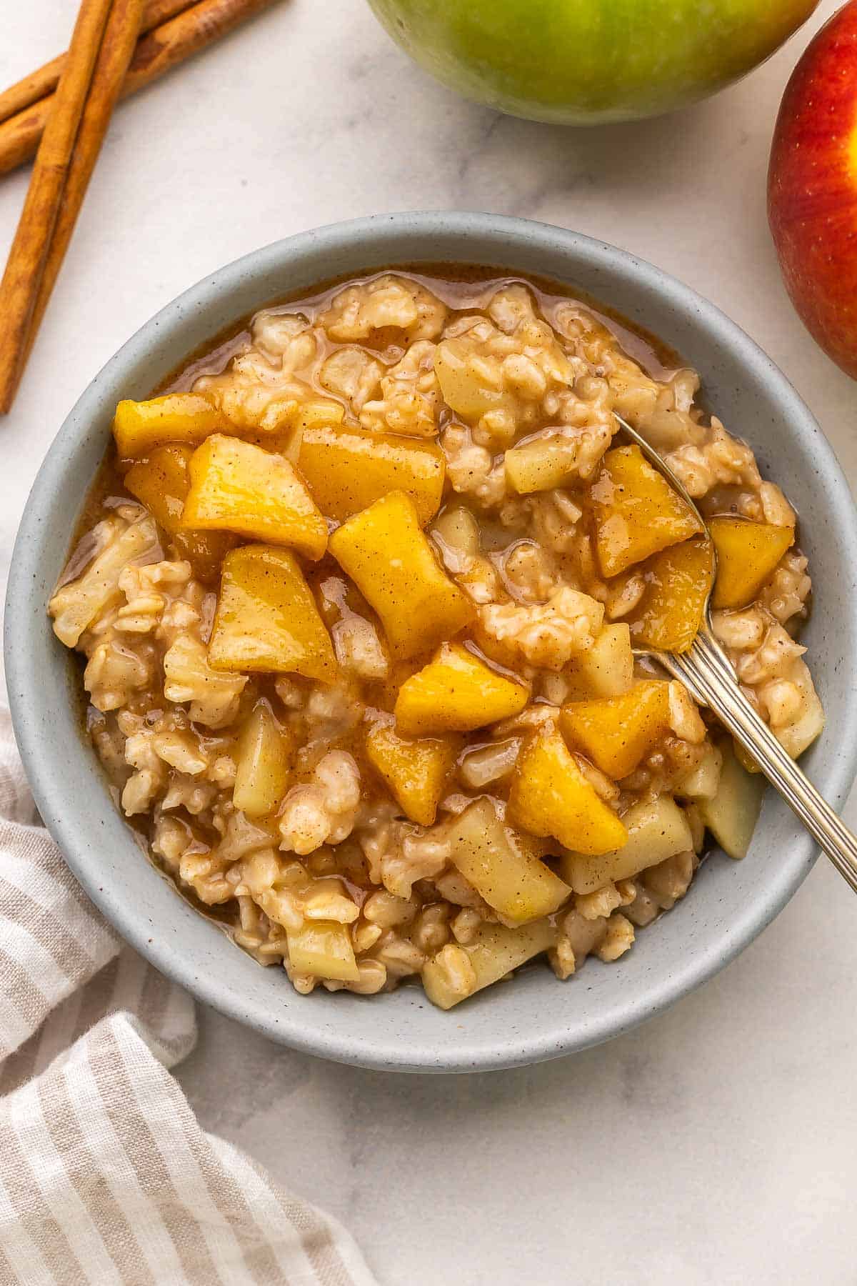This Apple Cinnamon Oatmeal recipe is packed with chunks of fresh apples swirled throughout warm creamy oats and topped with maple syrup. It's ready in 20 minutes and is gluten-free, dairy-free, and meal prep-friendly! #breakfast #recipe #apples | apple oatmeal | oatmeal recipe | how to cook oatmeal | apple recipes blue bowl of apple cinnamon oatmeal with a spoon stuck in.