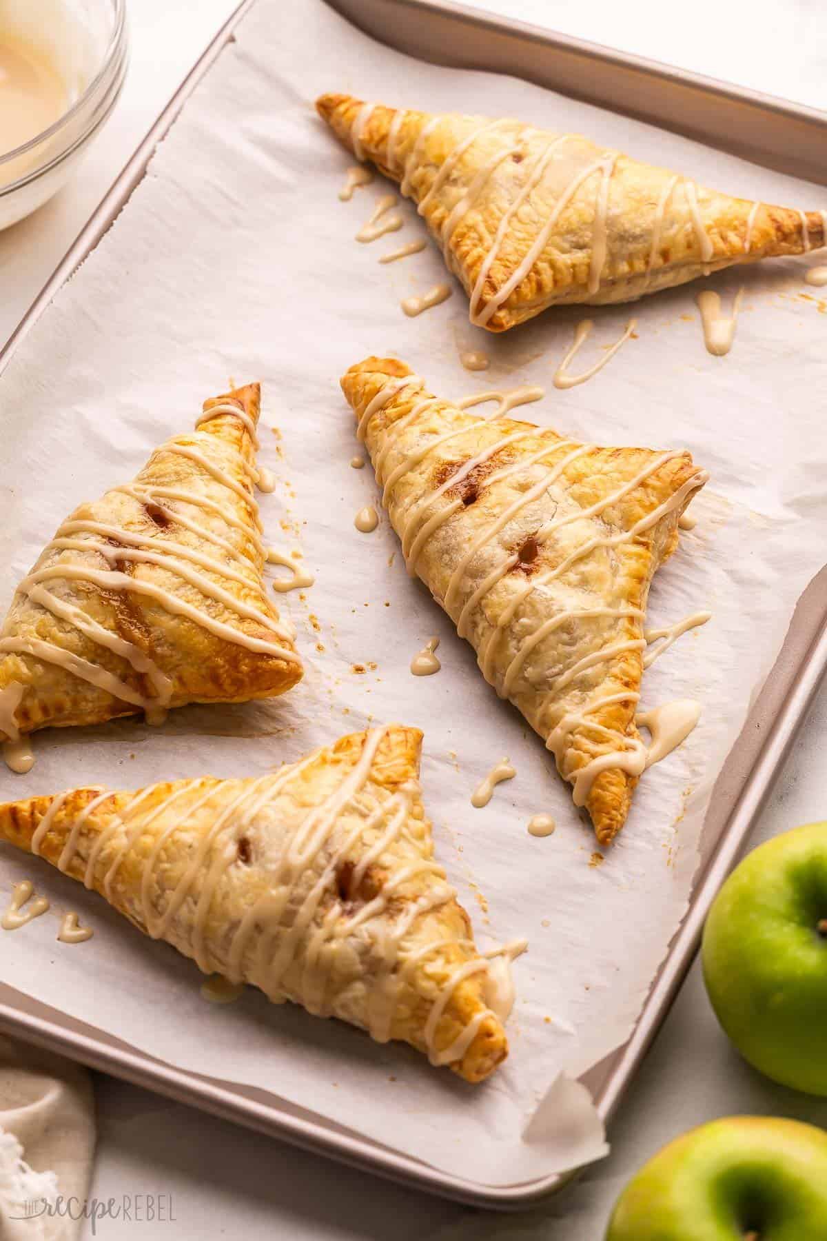These easy Apple Turnovers are filled with a sweet apple filling inside store-bought puff pastry sheets and drizzled with a luscious maple glaze. It's like apple pie wrapped up in a flaky, buttery pocket of goodness! #apple #dessert #recipe | apple recipes | apple dessert | fall baking | puff pastry recipes | apple cinnamon | sweet treat baked apple turnovers on a pan with apples beside.