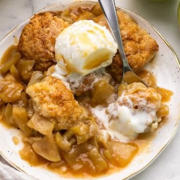 square image of caramel apple cobbler with ice cream and a spoon dug in.
