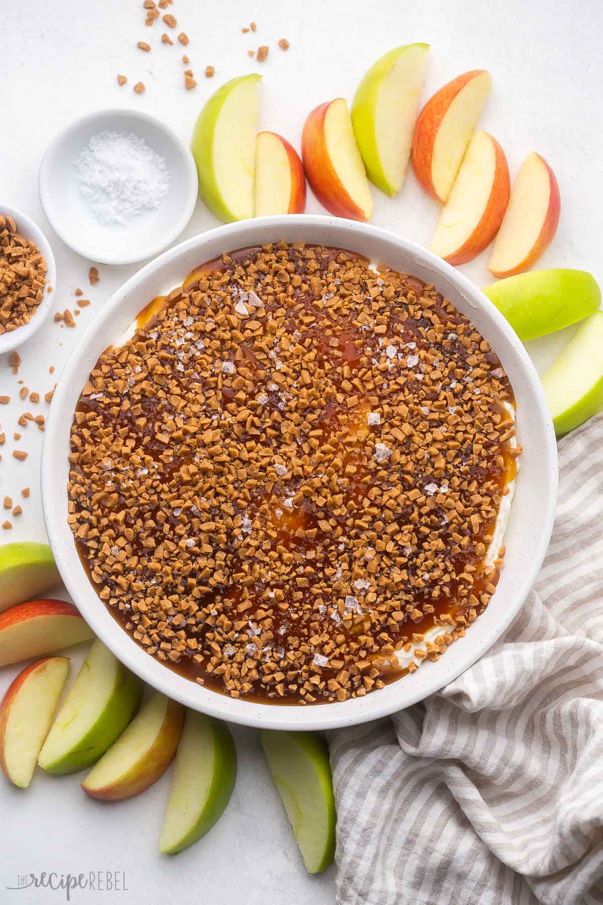 This easy Caramel Apple Dip recipe is the perfect dessert dip for fall, made with cream cheese and heavy whipping cream, with vanilla and caramel flavors, a crunchy caramel topping, and flaky sea salt! It's a crowd pleaser paired with crunchy apple slices. #dip #caramel #apple #recipes | fall recipes | apple recipes | caramel | salted caramel | caramel apple | dip recipe overhead view of a dish of caramel apple dip surrounded by sliced apples.