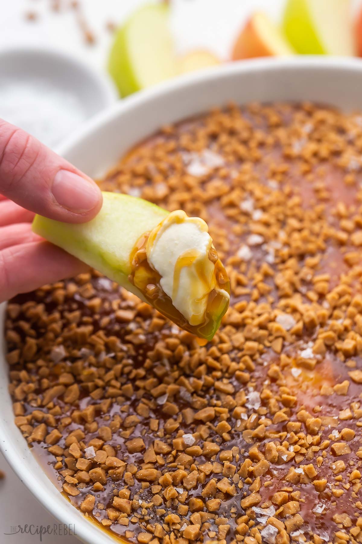 This easy Caramel Apple Dip recipe is the perfect dessert dip for fall, made with cream cheese and heavy whipping cream, with vanilla and caramel flavors, a crunchy caramel topping, and flaky sea salt! It's a crowd pleaser paired with crunchy apple slices. #dip #caramel #apple #recipes | fall recipes | apple recipes | caramel | salted caramel | caramel apple | dip recipe a hand holding an apple slice with caramel apple dip on it.