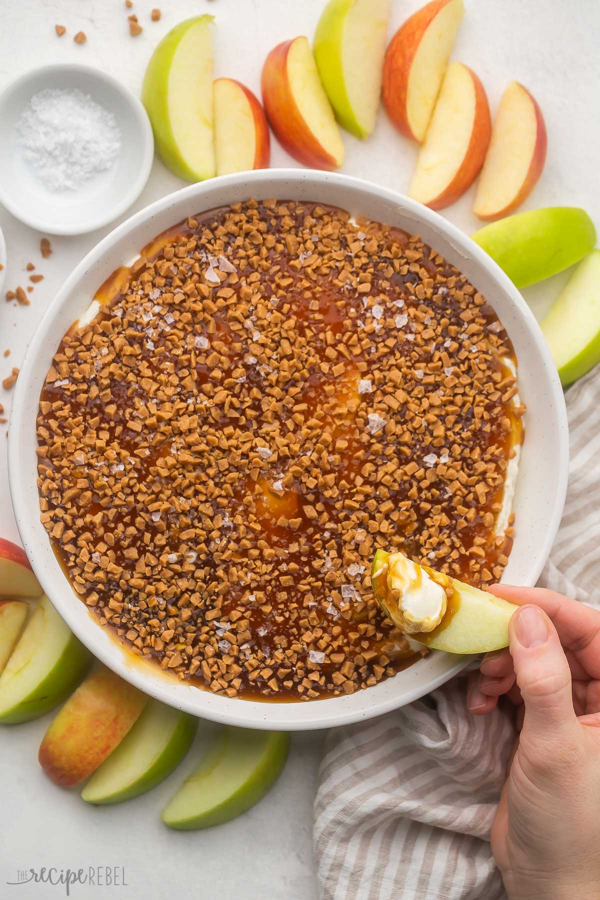 This easy Caramel Apple Dip recipe is the perfect dessert dip for fall, made with cream cheese and heavy whipping cream, with vanilla and caramel flavors, a crunchy caramel topping, and flaky sea salt! It's a crowd pleaser paired with crunchy apple slices. #dip #caramel #apple #recipes | fall recipes | apple recipes | caramel | salted caramel | caramel apple | dip recipe plate of caramel apple dip with apple slice held above.