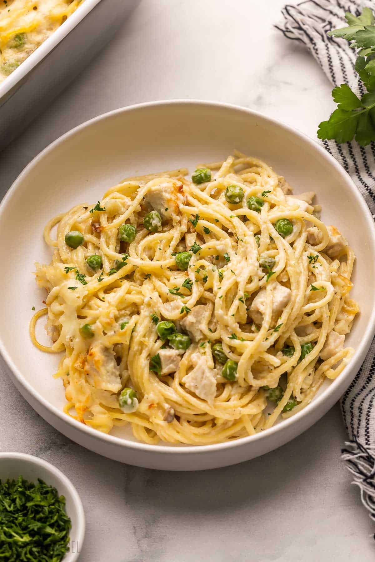 Chicken Tetrazzini is a cozy pasta bake recipe made with tender chicken, sautéed mushrooms, and peas, all tossed in a creamy homemade sauce, topped with mozzarella cheese and baked until golden! It's classic comfort food! #chicken #dinner #pasta | pasta bake | pasta recipe | casserole recipes | comfort food | leftover chicken | chicken pasta | easy dinner ideas | make ahead meal large bowl filled with chicken tetrazzini and parsley beside.