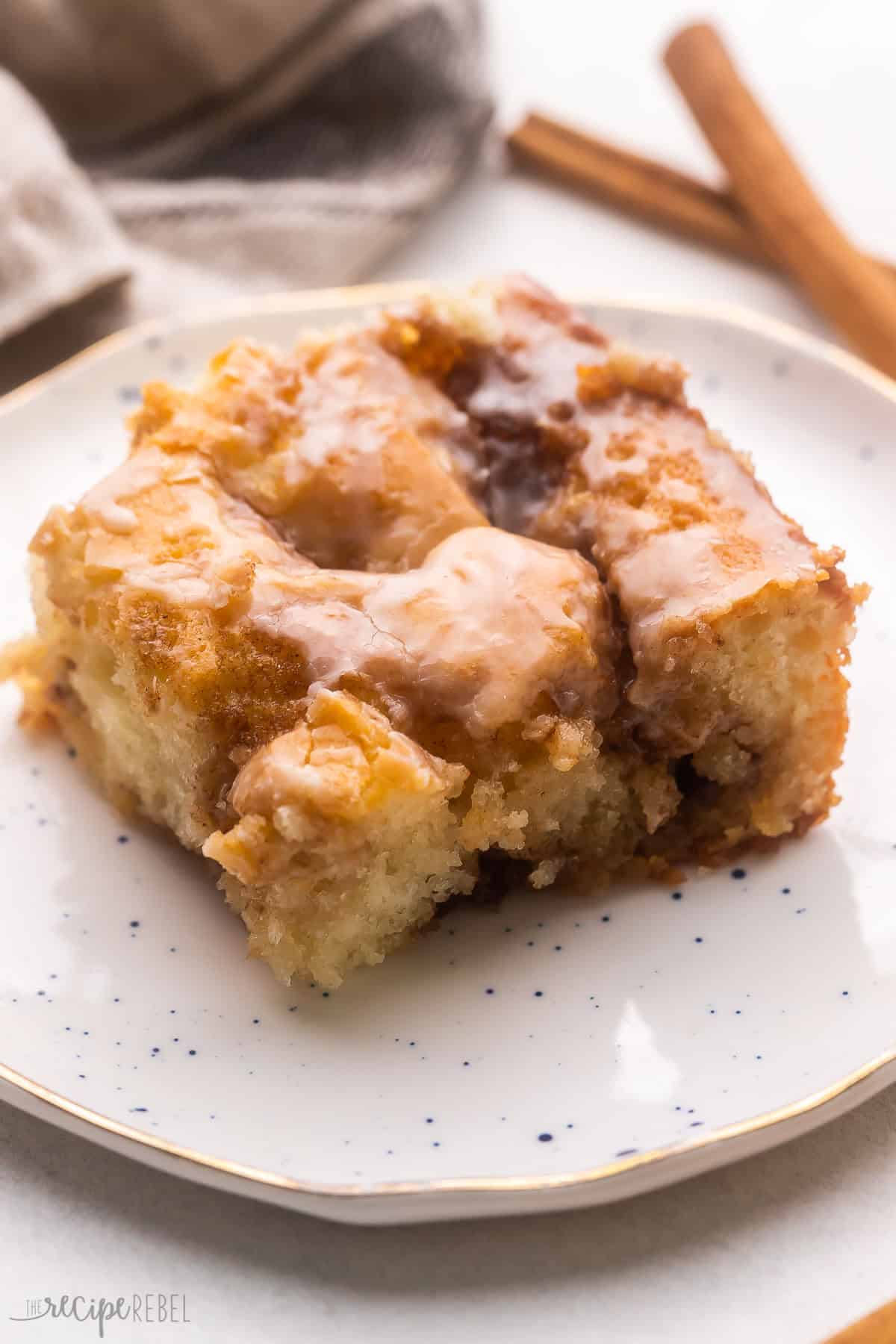 Cinnamon Roll Cake feature a soft homemade cake that's swirled with the most scrumptious cinnamon roll filling! It's baked to perfection in a casserole dish in just 30 minutes before being served with a generous amount of sugary sweet glaze. #cake #dessert #recipe | dessert recipes | cake recipes | cinnamon recipes | brunch | christmas breakfast | christmas dessert | cinnamon roll recipes | thanksgiving breakfast a piece of cinnamon roll cake on a white plate with cinnamon sticks behind it.