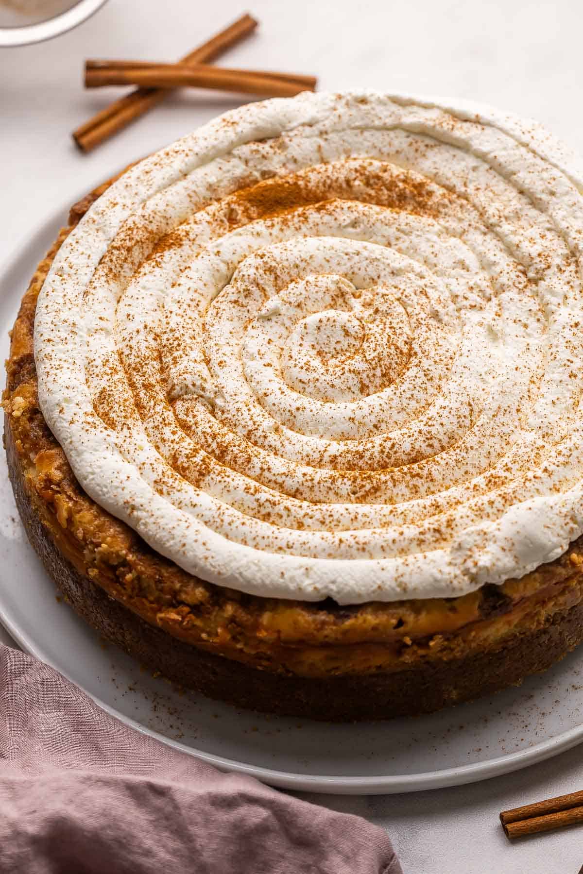 This Cinnamon Roll Cheesecake is made with a buttery cinnamon sugar filling between layers of rich, creamy cheesecake in a buttery graham cracker crust. No water bath and no cracks! | cheesecake recipe | cinnamon bun cheesecake | cinnamon rolls | cinnamon desserts | cinnamon recipes | holiday desserts | dessert recipe a full cinnamon roll cheesecake on a white plate with cinnamon sticks behind it.