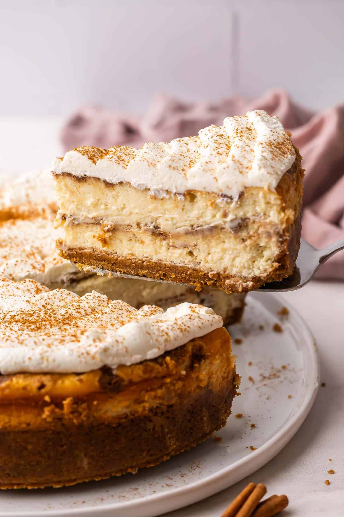 This Cinnamon Roll Cheesecake is made with a buttery cinnamon sugar filling between layers of rich, creamy cheesecake in a buttery graham cracker crust. No water bath and no cracks! | cheesecake recipe | cinnamon bun cheesecake | cinnamon rolls | cinnamon desserts | cinnamon recipes | holiday desserts | dessert recipe a slice of cheesecake being lifted off of plate.