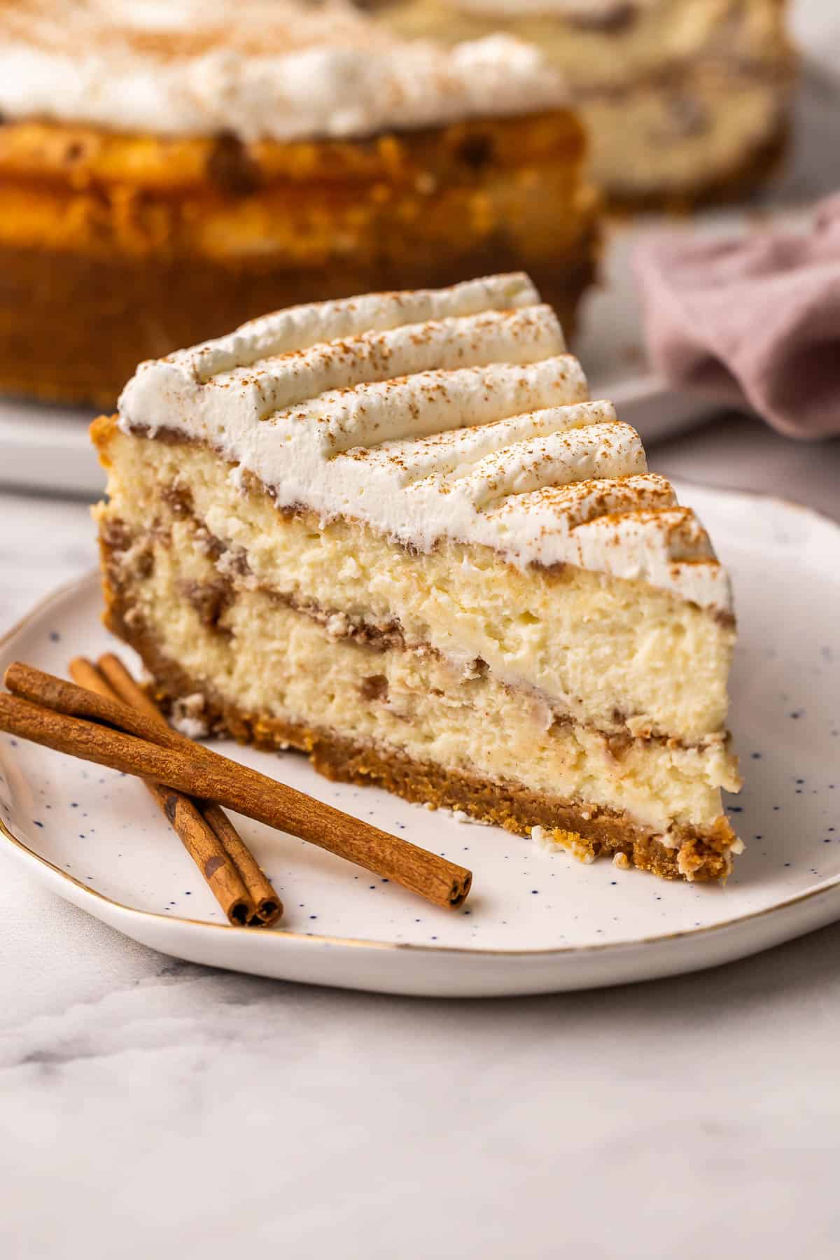 This Cinnamon Roll Cheesecake is made with a buttery cinnamon sugar filling between layers of rich, creamy cheesecake in a buttery graham cracker crust. No water bath and no cracks! | cheesecake recipe | cinnamon bun cheesecake | cinnamon rolls | cinnamon desserts | cinnamon recipes | holiday desserts | dessert recipe a slice of cinnamon roll cheesecake on a speckled plate with cinnamon sticks beside.