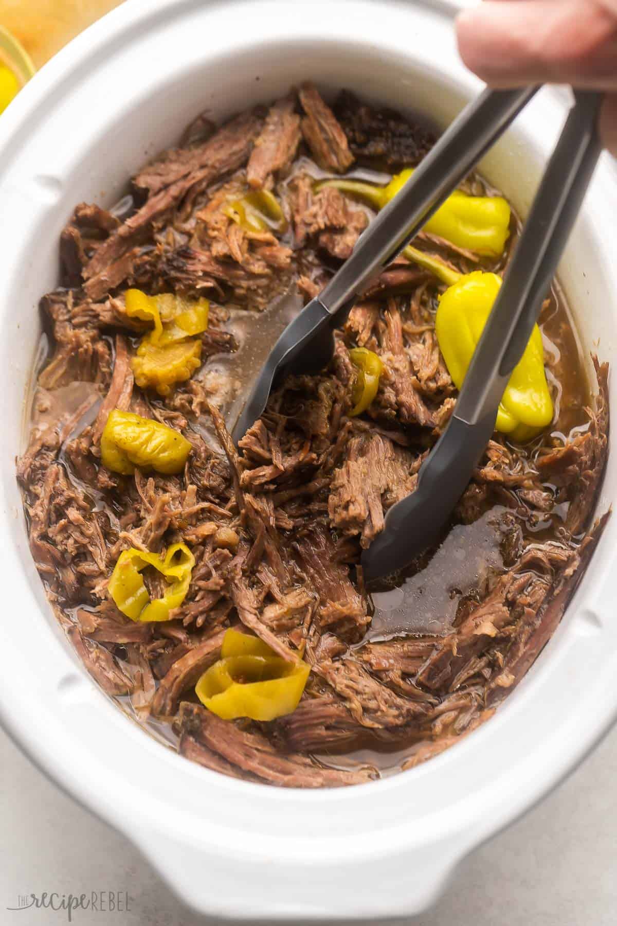 Easy shredded Italian Beef made in the crockpot, just six ingredients! Tender chuck roast is simmered in a tangy broth for fall-apart goodness, perfect for serving on sandwiches. Includes Instant Pot method and step-by-step video. #slowcooker #crockpot #recipe #dinner | beef roast recipe | beef recipe | roast beef | mississippi beef | sandwich recipe | crockpot dinner tongs lifting italian beef out of white crockpot.