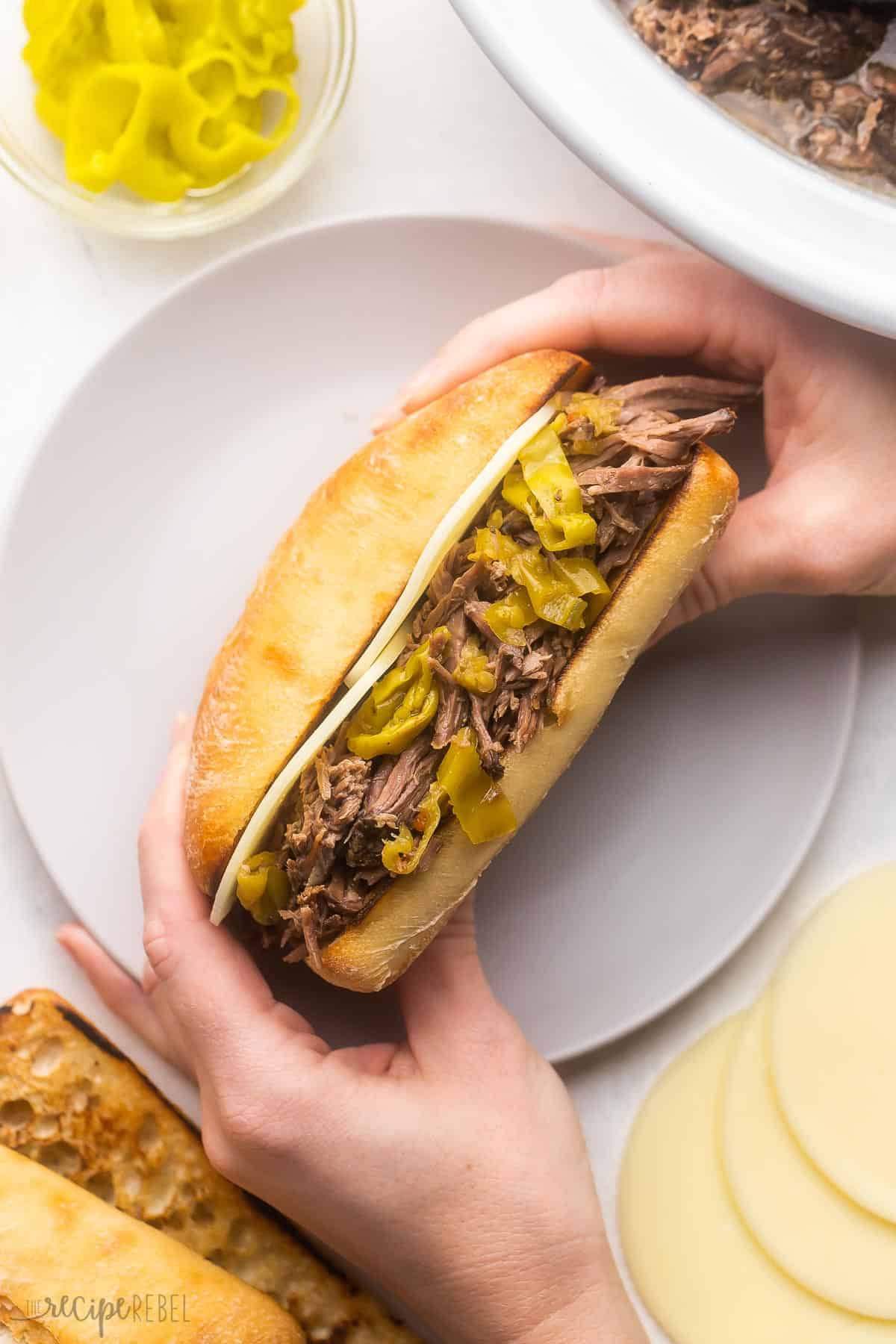 Easy shredded Italian Beef made in the crockpot, just six ingredients! Tender chuck roast is simmered in a tangy broth for fall-apart goodness, perfect for serving on sandwiches. Includes Instant Pot method and step-by-step video. #slowcooker #crockpot #recipe #dinner | beef roast recipe | beef recipe | roast beef | mississippi beef | sandwich recipe | crockpot dinner hands holding a crockpot italian beef sandwich over a plate.