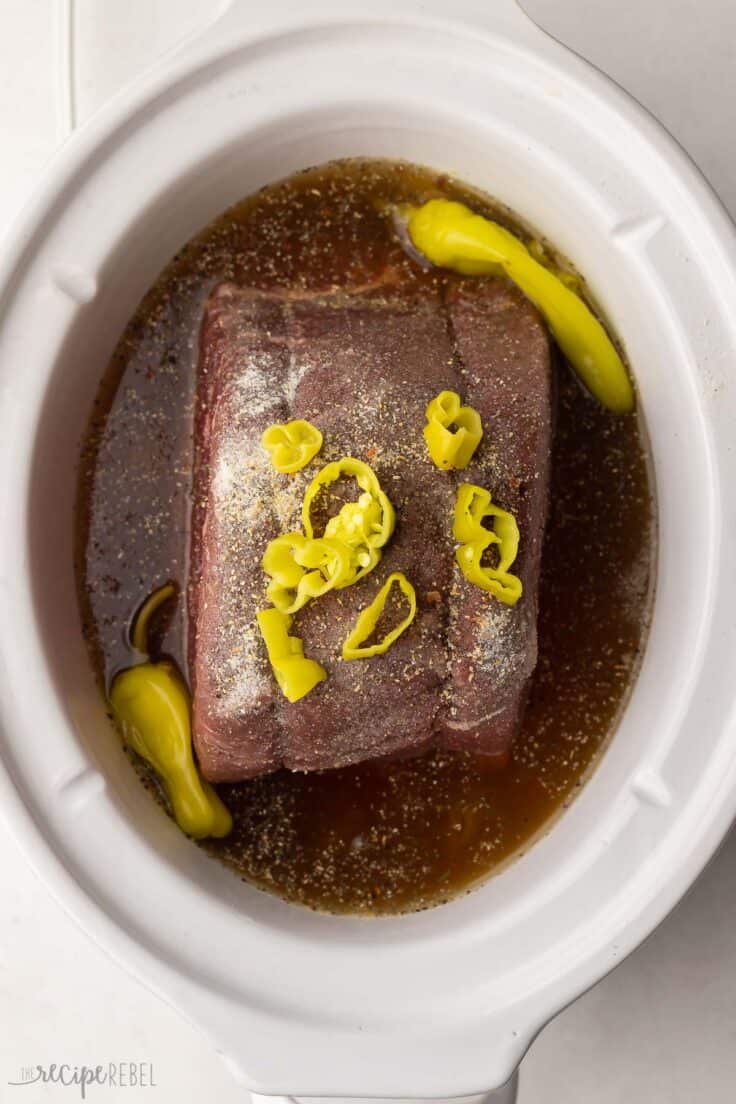beef roast in white crockpot with pepperoncini peppers on top.