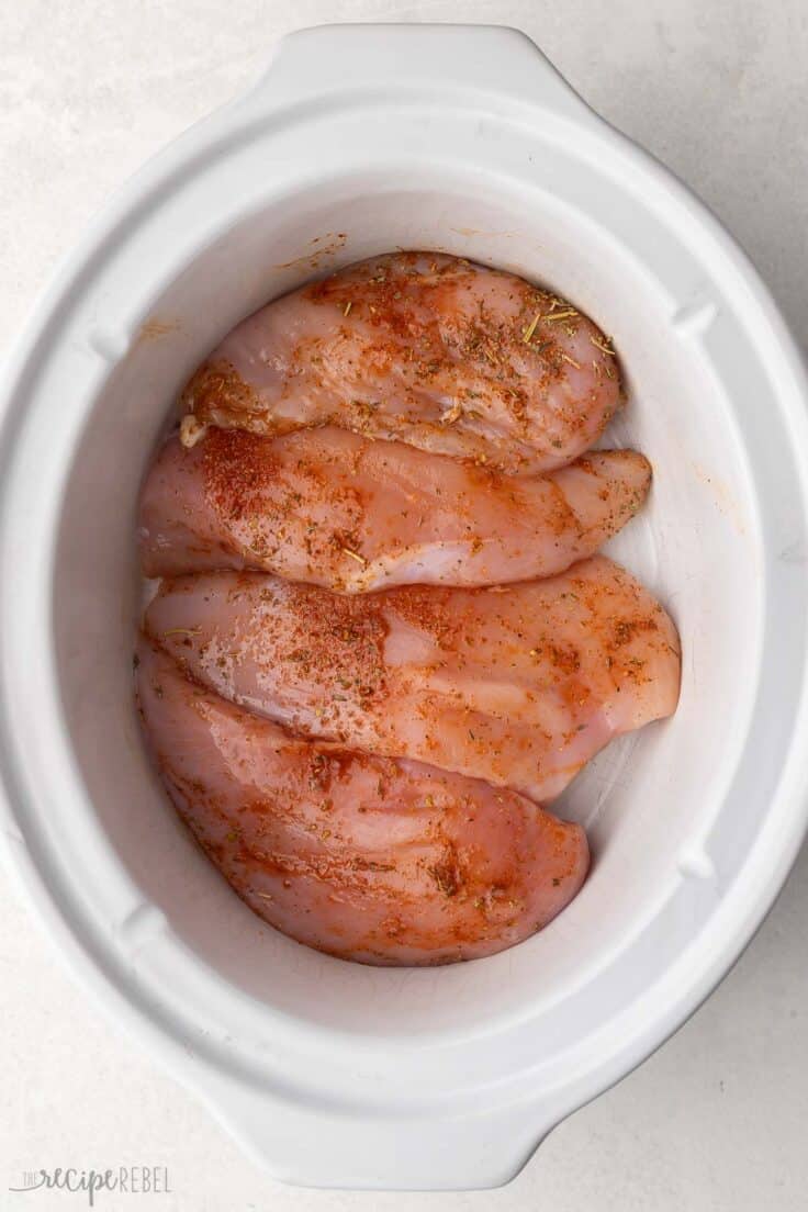 four uncooked seasoned chicken breasts in a white crockpot.