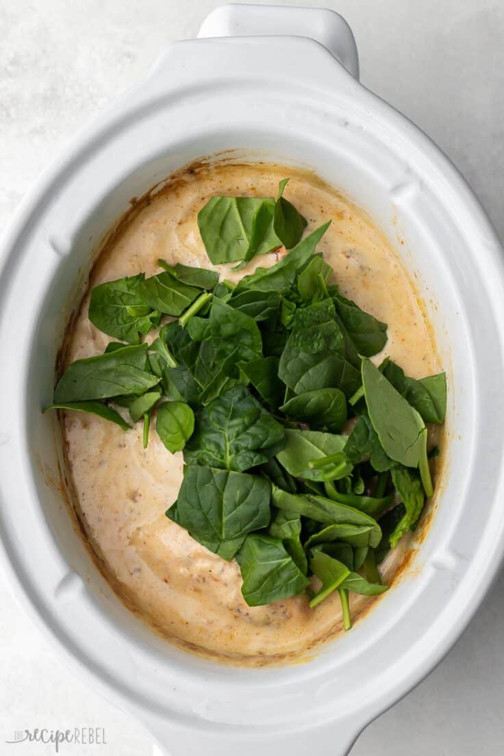 white slow cooker with spinach added to top of other ingredients.