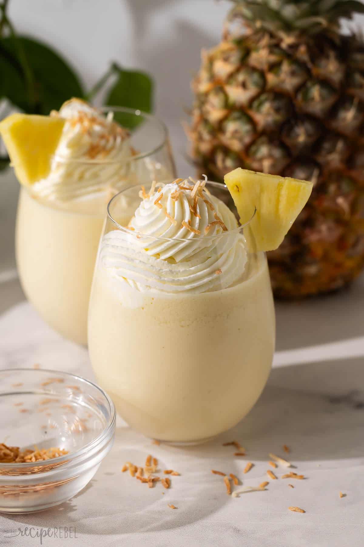 Get a taste of the tropics with this Frozen Pina Colada slushy recipe! It's made with creamy coconut milk, condensed milk, vanilla, frozen pineapple, and tons of ice for a drink that feels like a beach vacation. #drink #recipe #mocktail | mocktail recipe | drink recipe | summer drink | frozen drinks | blended drink recipe two glasses of pina colada slushy topped with whipped cream.
