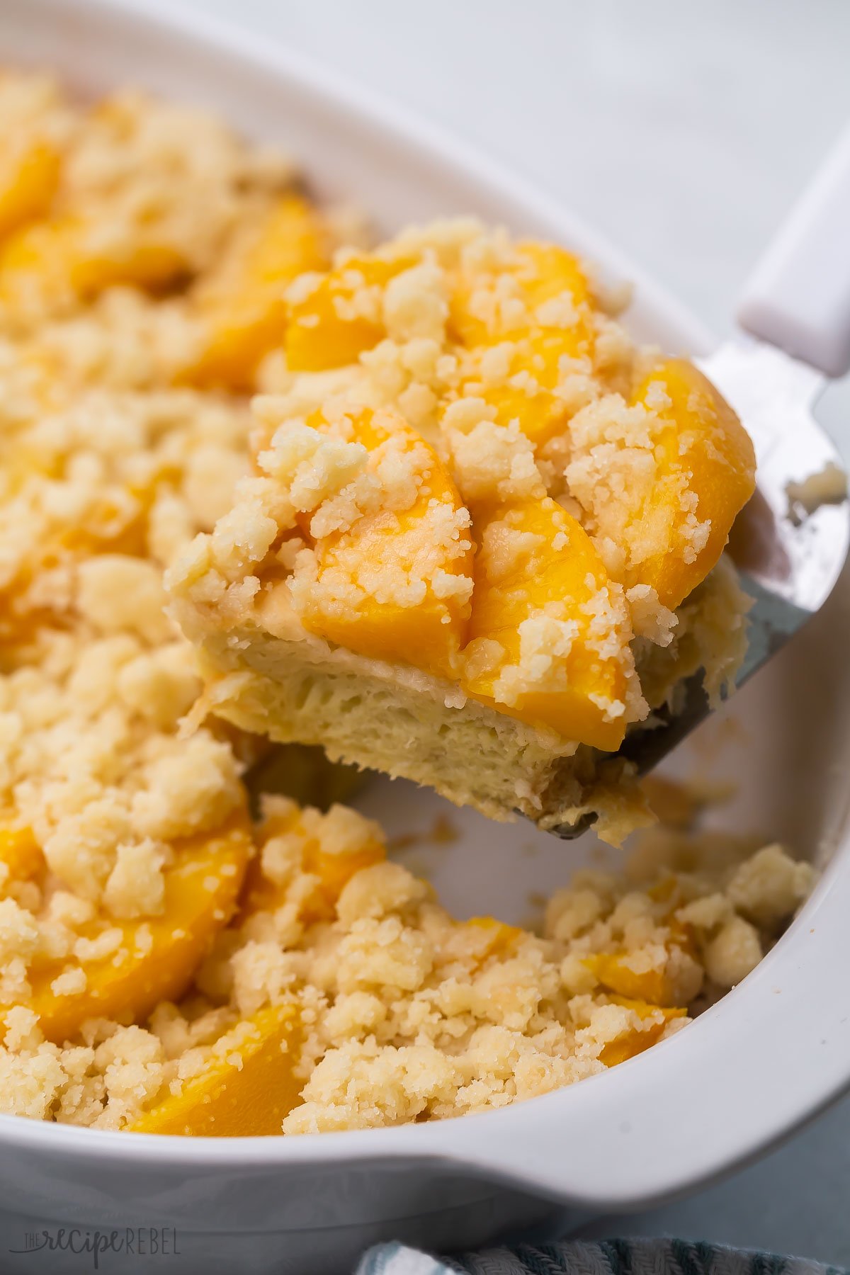 An easy Overnight French Toast Casserole topped with peach cobbler — it's the BEST French toast I’ve ever had! So perfect for Spring, it's full of juicy peaches and a sweet, moist crumb topping! #peaches #recipe | breakfast recipe | brunch ideas | easter brunch | canned peaches | fresh peaches | french toast casserole Close up of a slice of peach cobbler french toast being lifted out of a casserole dish.