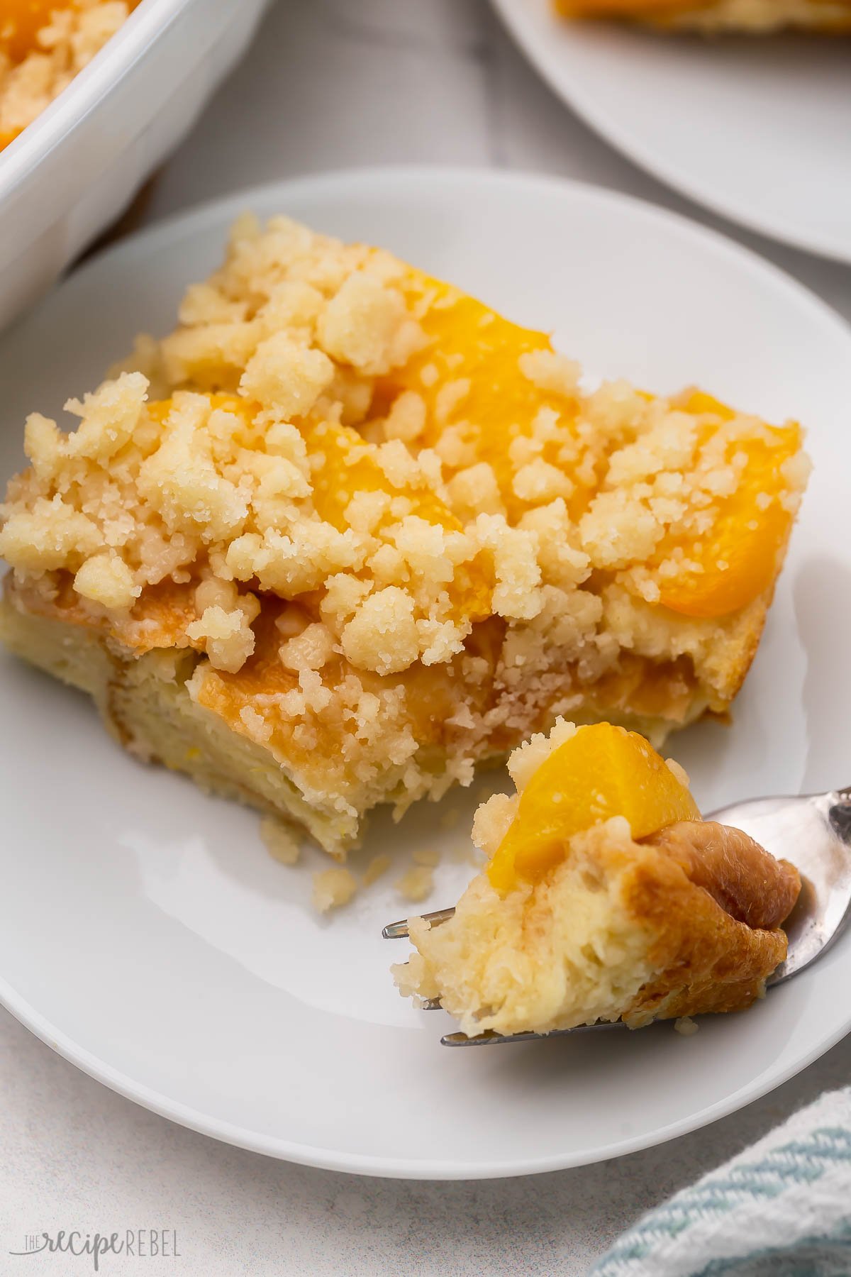 An easy Overnight French Toast Casserole topped with peach cobbler — it's the BEST French toast I’ve ever had! So perfect for Spring, it's full of juicy peaches and a sweet, moist crumb topping! #peaches #recipe | breakfast recipe | brunch ideas | easter brunch | canned peaches | fresh peaches | french toast casserole Close up of a slice of peach cobbler French toast on a white plate with a fork having taken a forkful of it.