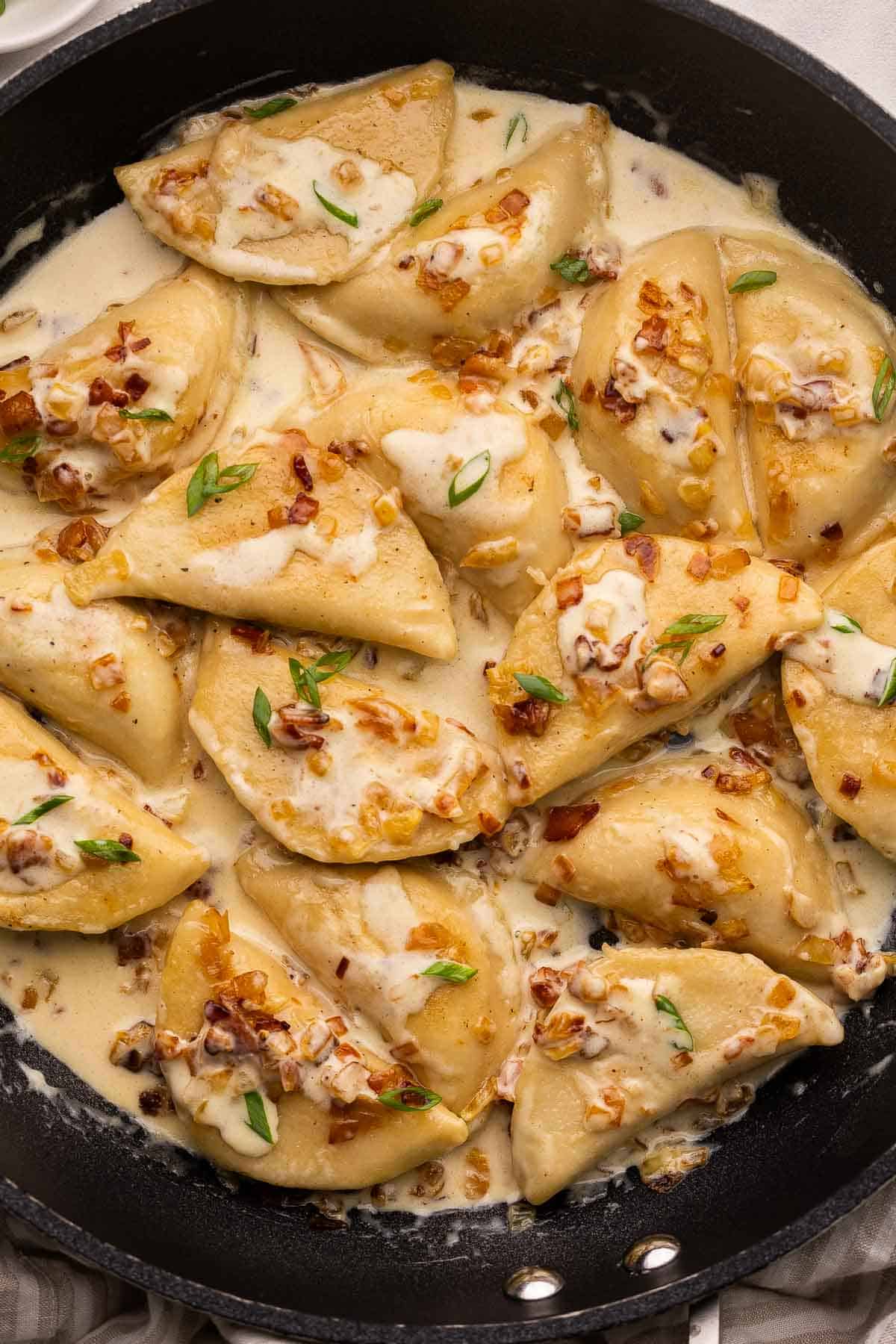 This Perogies recipe with Bacon and Onions recipe is made with frozen perogies swimming in a creamy sauce with smoky bacon and onions. Ready in 35 minutes, it's the perfect side dish for the holidays or whenever you're craving something comforting. | dinner | recipe | easy dinner ideas | pierogi | comfort food | skillet meals perogies covered in a creamy sauce in a large black pan.