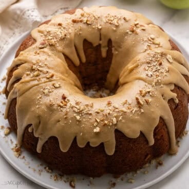 a full apple bundt cake on a white plate topped with chopped pecans.