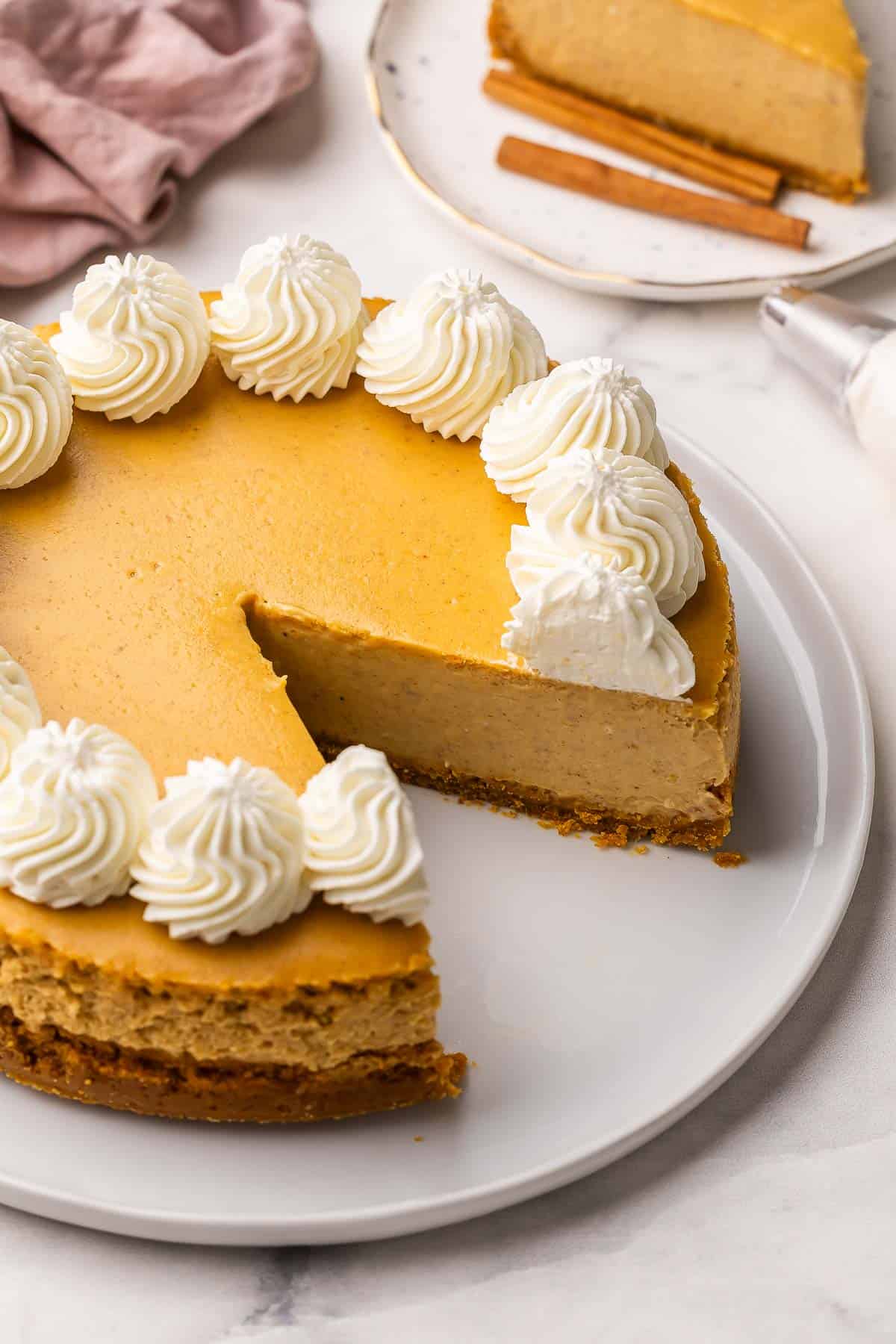 This baked Pumpkin Cheesecake has a super creamy filling and is packed with warm spices and cozy pumpkin flavor! It's easier than the rest, thanks to one secret method. No water bath and no cracks! #pumpkin #cheesecake #dessert | dessert recipes | pumpkin recipes | pumpkin dessert | baked cheesecake recipe | fall dessert | thanksgiving dessert pumpkin cheesecake on a white plate with a piece missing.