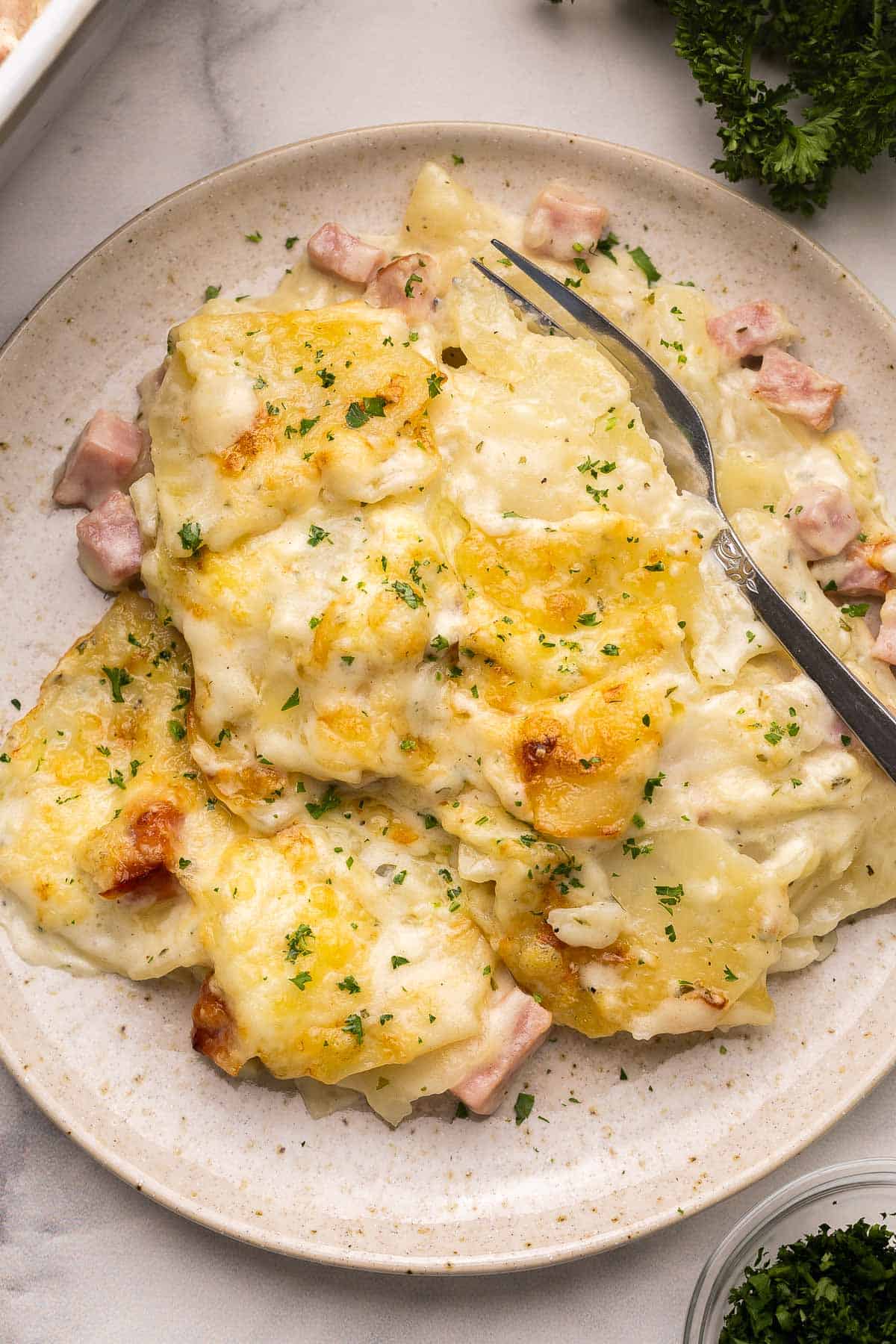 Scalloped Potatoes and Ham is a cheesy, creamy potato casserole made with layers of potatoes and ham cooked in a creamy white sauce. It's a comforting side dish perfect for a holiday meal or Sunday dinner! #dinner #recipe | potatoes | potato recipe | casserole recipe | holiday side dish | side dishes | comfort food a plate filled with scalloped potatoes and ham with parsley behind it.