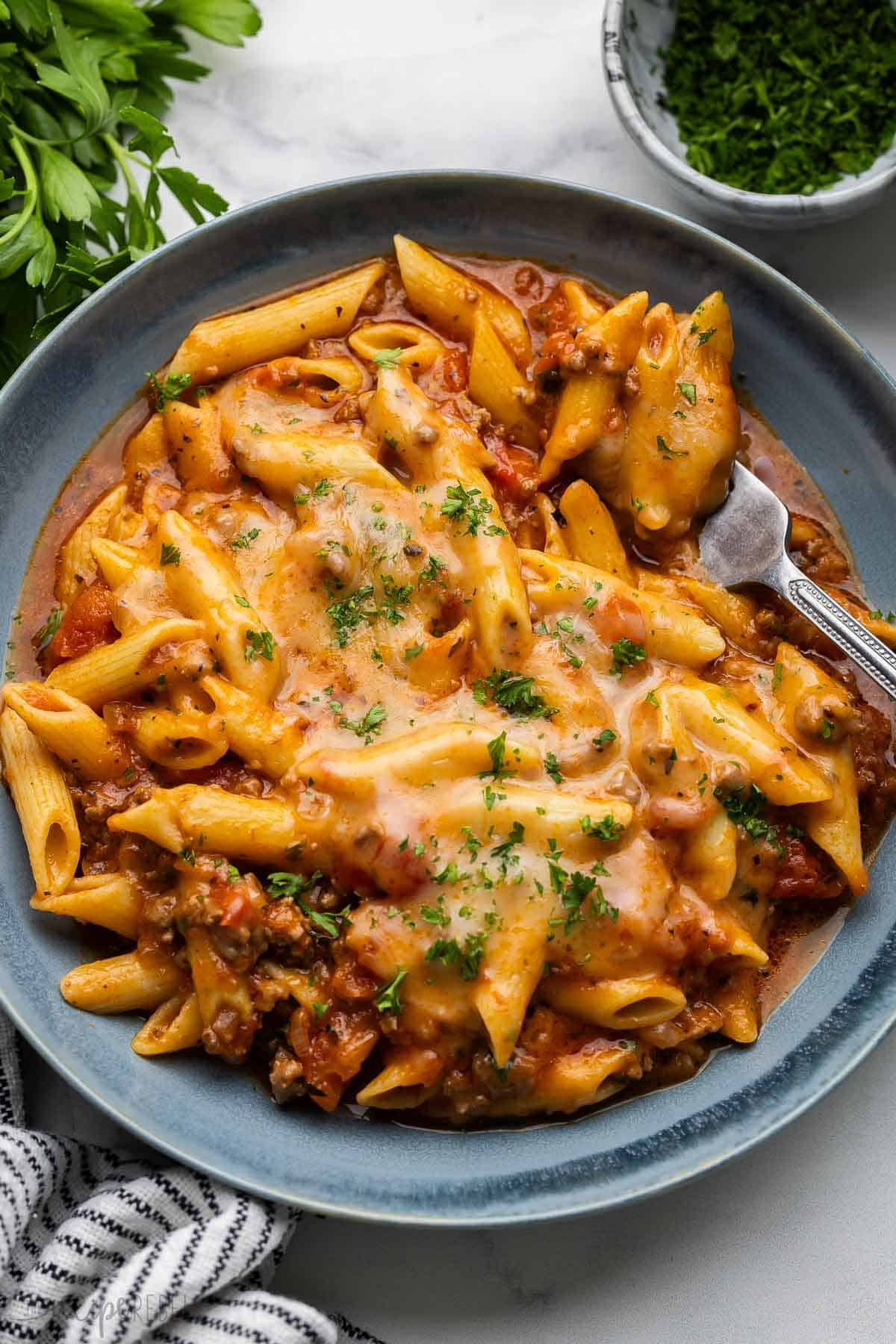 This Slow-Cooker Baked Ziti is made completely in the crockpot—even the pasta! It is so much easier than the traditional version but packs all of the same flavor! The recipe includes a step-by-step video. #recipe #dinner #slowcooker | slow cooker dinner | crockpot meals | crockpot recipes | slow cooker recipes | ground beef recipes | pasta recipe | easy dinner ideas | ground beef pasta blue plate filled with baked ziti and topped with chopped parsley.