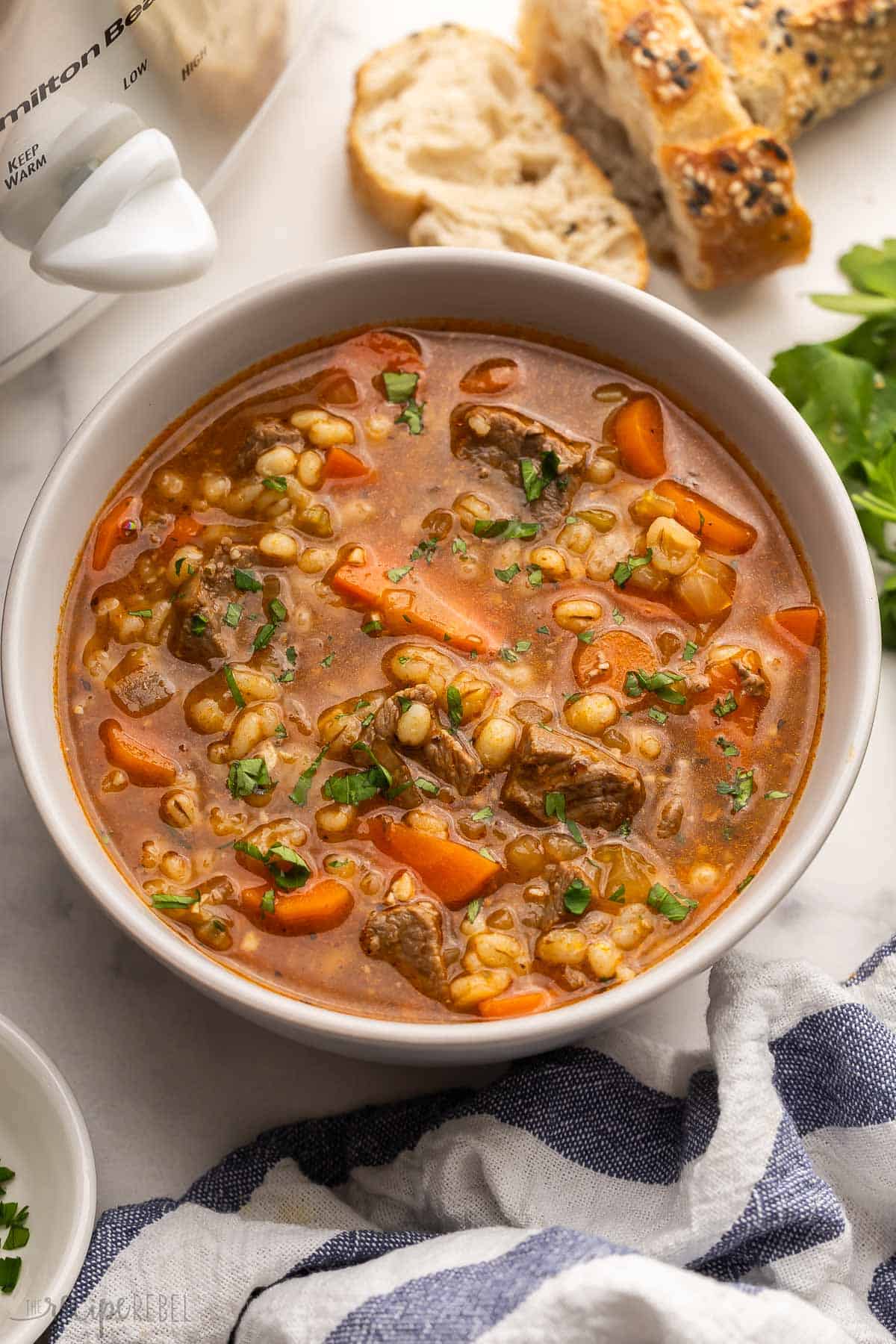 This Slow Cooker Beef Barley Soup is loaded with tender beef, barley, and vegetables simmering in a rich broth. It's the perfect crockpot meal for warming you up on a chilly day! #soup #recipe #dinner | soup recipes | beef recipes | comfort food | slow cooker soup | crockpot soups | crockpot meals | easy dinner ideas large bowl full of beef barley soup with bread in the background.