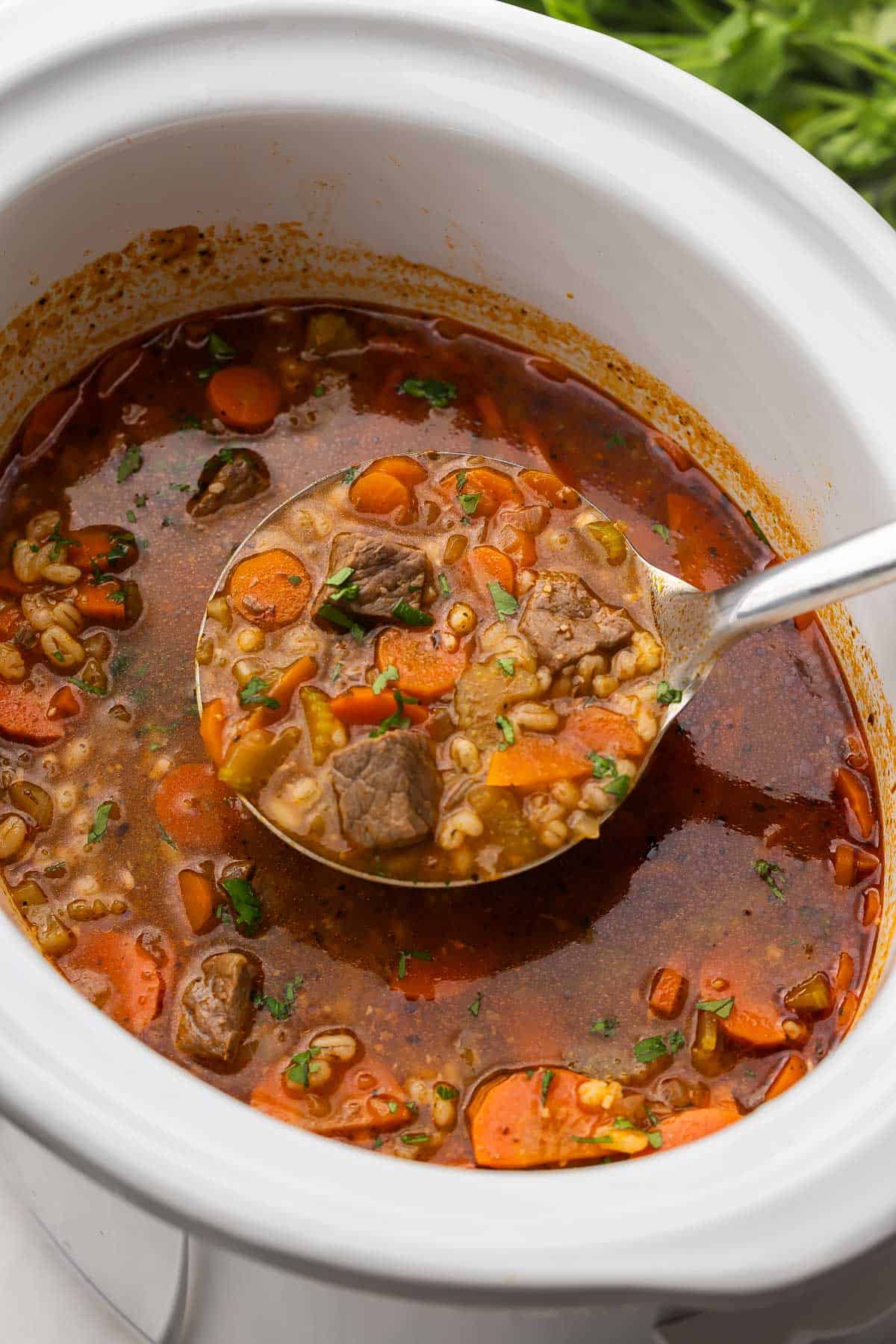 This Slow Cooker Beef Barley Soup is loaded with tender beef, barley, and vegetables simmering in a rich broth. It's the perfect crockpot meal for warming you up on a chilly day! #soup #recipe #dinner | soup recipes | beef recipes | comfort food | slow cooker soup | crockpot soups | crockpot meals | easy dinner ideas close up image of a metal ladle scooping slow cooker beef barley soup.