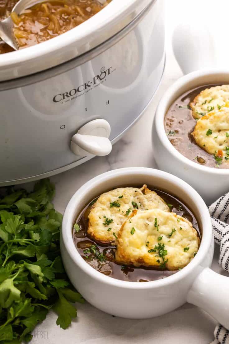 two bowls of french onion soup beside white slow cooker.