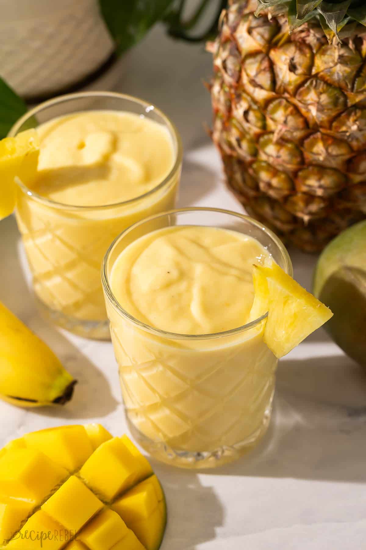 This Tropical Smoothie is made with banana, mango, pineapple, Greek yogurt, and coconut milk or pineapple juice! It's a great way to enjoy a taste of the tropics without leaving your kitchen. #smoothie #recipe #drink | drink recipes | smoothie recipes | fruit smoothies | healthy recipes | breakfast | healthy snack two glasses of tropical smoothie with a pineapple slice on the side.